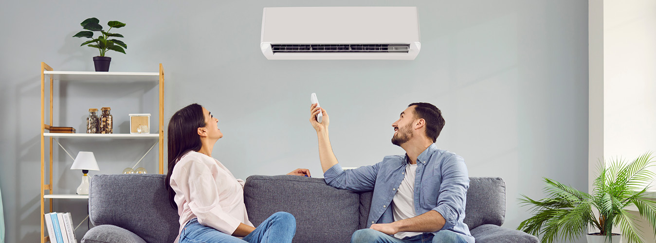 Young happy man and woman turning on air conditioner sitting on sofa at home. Smiling couple of homeowners enjoying cool conditioned air using remote resting on couch together in living room.